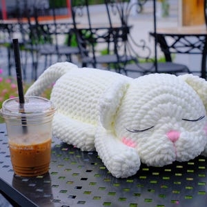 May include: A handmade, white crochet bunny plushie with closed eyes and pink accents, resting on a metal table. A clear plastic cup of iced coffee with a black straw sits nearby. The bunny is the focal point.