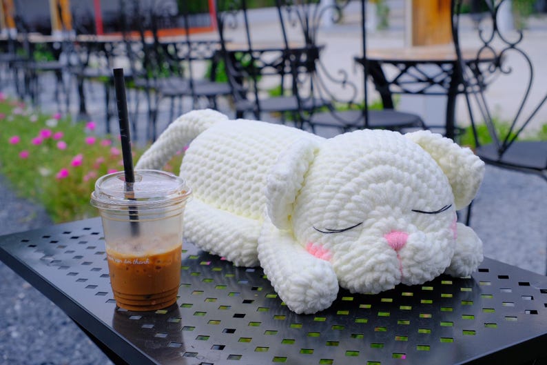 May include: A handmade, white crochet bunny plushie with closed eyes and pink accents, resting on a metal table. A clear plastic cup of iced coffee with a black straw sits nearby. The bunny is the focal point.