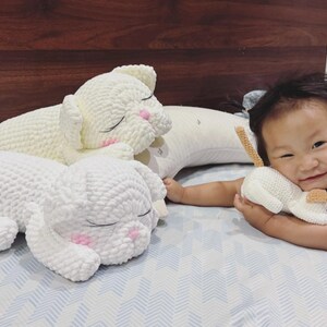 May include: Three white crochet cat plushies. Two large plushies are lying down with closed eyes and pink cheeks. A smaller plushie is held by a person. The plushies have a textured, soft appearance.