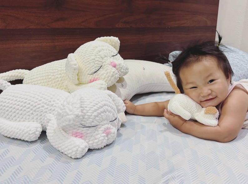 May include: Three white crochet cat plushies. Two large plushies are lying down with closed eyes and pink cheeks. A smaller plushie is held by a person. The plushies have a textured, soft appearance.