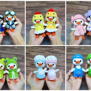 Op de afbeelding: Een collectie van twaalf kleine, handgemaakte gehaakte eendenpoppen, elk met verschillende outfits zoals vliegersuitrusting, gele kuikenkostuums, roze en paarse hoeden, groene kikkercapuchons, koksmutsen en blauwe of groene tuinbroeken met bijpassende petten.