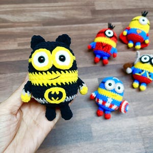 May include: A collection of handmade crochet Minion dolls, each with unique superhero-themed designs. The dolls feature vibrant colors like yellow, blue, red, and black, with details such as capes and emblems. The dolls are approximately 4 inches tall.