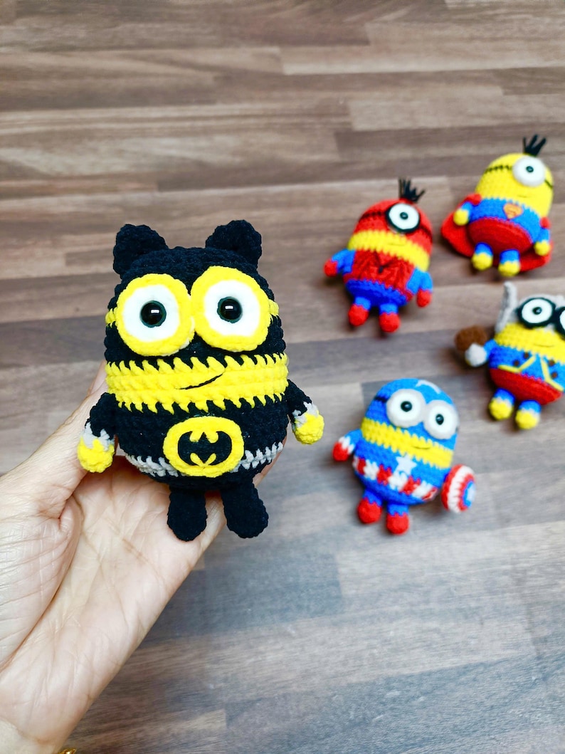 May include: A collection of handmade crochet Minion dolls, each with unique superhero-themed designs. The dolls feature vibrant colors like yellow, blue, red, and black, with details such as capes and emblems. The dolls are approximately 4 inches tall.