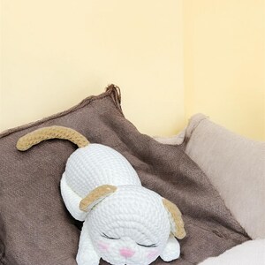 May include: A white crocheted dog toy with tan ears and tail, lying on a brown pillow. The dog has closed eyes and pink cheeks. The background is a light yellow wall and a beige pillow.