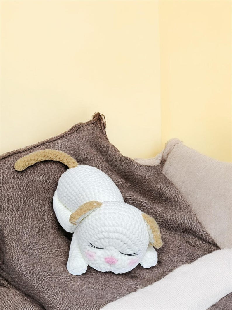 May include: A white crocheted dog toy with tan ears and tail, lying on a brown pillow. The dog has closed eyes and pink cheeks. The background is a light yellow wall and a beige pillow.