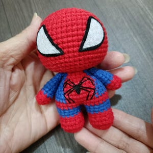 May include: A small crocheted stuffed toy of Spiderman. The toy is red and blue with a black spider design on the chest. The toy has white eyes with black outlines.