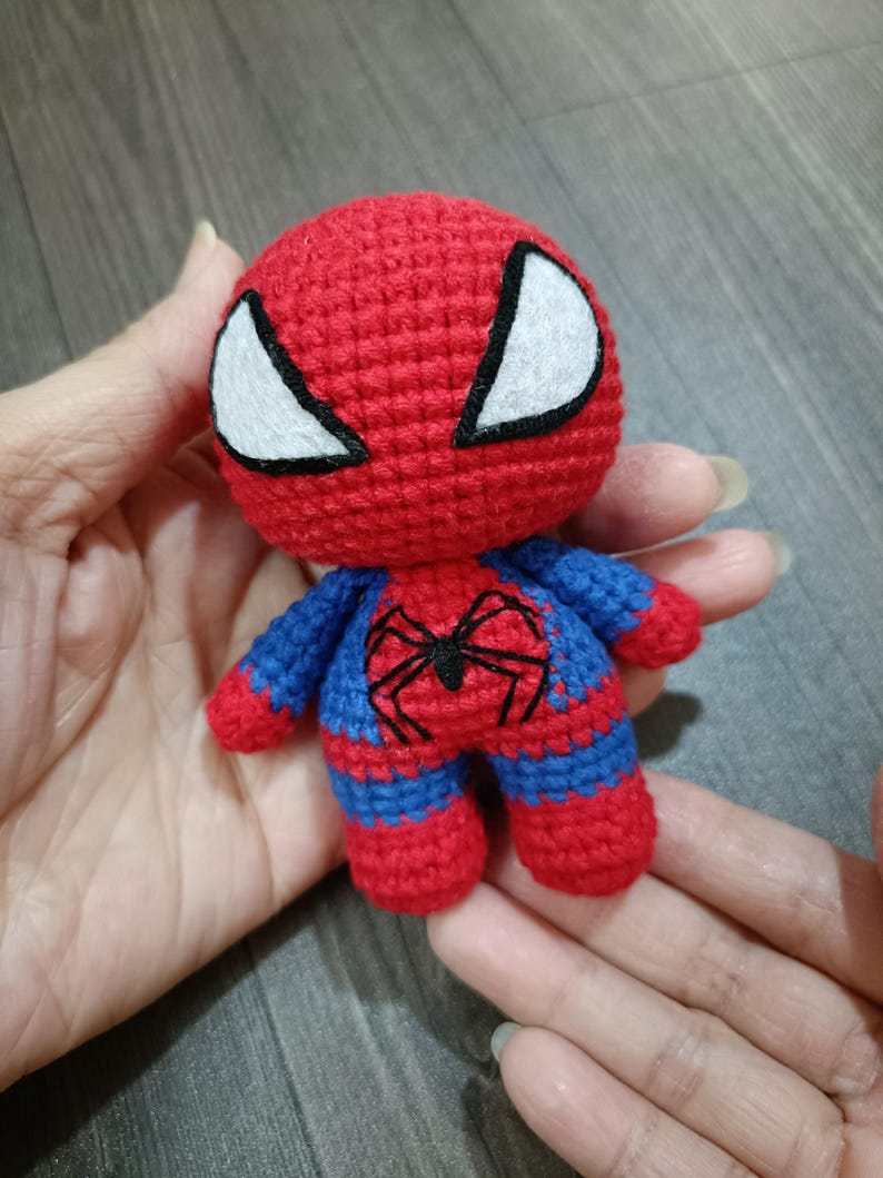 May include: A small crocheted stuffed toy of Spiderman. The toy is red and blue with a black spider design on the chest. The toy has white eyes with black outlines.