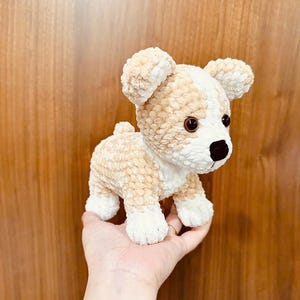 May include: A crocheted stuffed dog toy with brown and white fur. The dog is standing on its four paws and has a black nose and brown eyes.