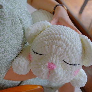 May include: A white, crocheted bunny plushie with closed eyes and pink accents on the nose and cheeks. The bunny is soft and fluffy, with large ears and a rounded body. The plushie is being held.