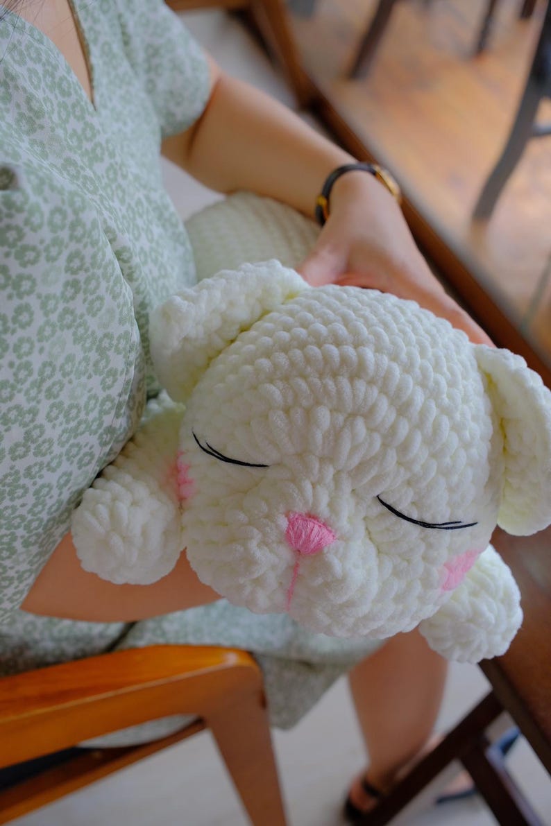 May include: A white, crocheted bunny plushie with closed eyes and pink accents on the nose and cheeks. The bunny is soft and fluffy, with large ears and a rounded body. The plushie is being held.