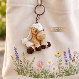 May include: A cream-coloured tote bag with an embroidered floral design including lavender and daisies. A crocheted horse keyring with a white mane and brown details hangs from the bag.