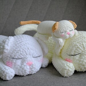 May include: Three white crochet cat pillows with closed eyes and pink accents. One small cat pillow rests on a larger one. The pillows are soft and fluffy, perfect for a nursery or child's room.