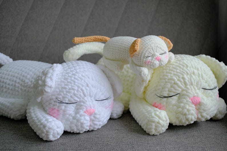 May include: Three white crochet cat pillows with closed eyes and pink accents. One small cat pillow rests on a larger one. The pillows are soft and fluffy, perfect for a nursery or child's room.