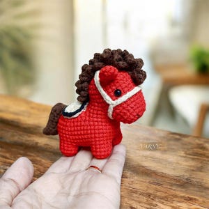 Puede incluir: Un caballo rojo hecho a mano con ganchillo, con crin y cola marrones. El caballo tiene una brida blanca y una pequeña silla de montar. El juguete se sostiene en una mano, mostrando el tamaño y los detalles de la artesanía.