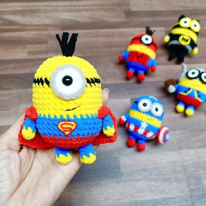 May include: A collection of handmade crochet Minion-inspired figures. The central figure is yellow with a single eye, wearing a red cape and a blue and red outfit with a Superman logo. Other figures include a bee and Captain America-themed Minions.