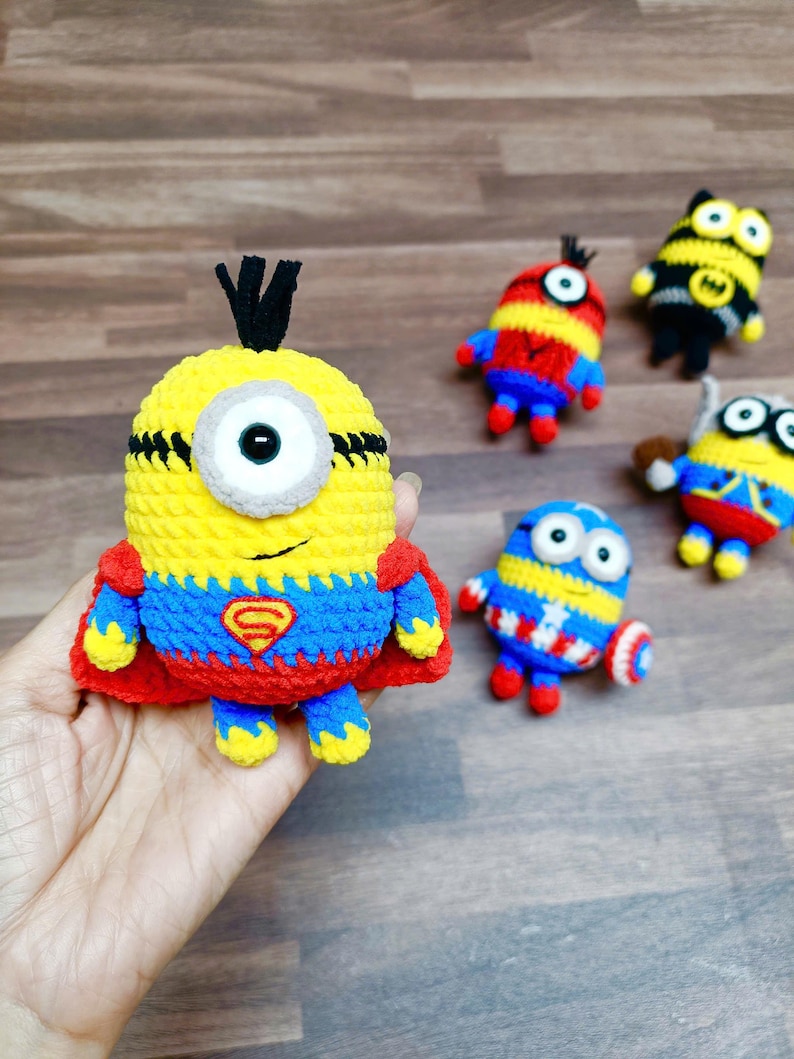 May include: A collection of handmade crochet Minion-inspired figures. The central figure is yellow with a single eye, wearing a red cape and a blue and red outfit with a Superman logo. Other figures include a bee and Captain America-themed Minions.