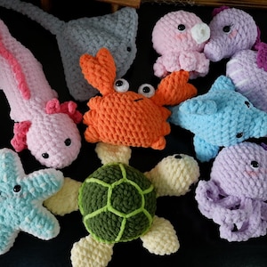Puede incluir: Una colección de peluches de criaturas marinas de crochet, que incluyen una estrella de mar, una tortuga, un cangrejo, un pulpo, un ajolote, un caballito de mar y dos rayas. Los peluches están hechos con varios colores de hilo, incluyendo azul, verde, naranja, rosa y morado.