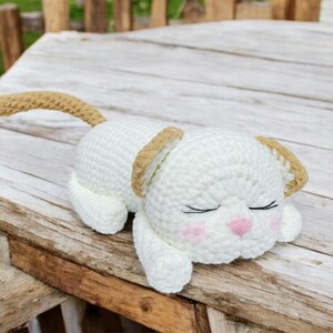 May include: A handmade, white crochet cat toy with tan accents on the ears and tail. The cat is lying down with closed eyes, pink cheeks, and a pink nose. The toy is on a wooden surface.