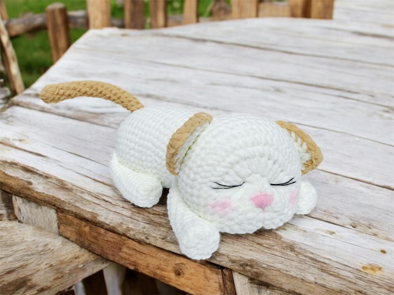 May include: A handmade, white crochet cat toy with tan accents on the ears and tail. The cat is lying down with closed eyes, pink cheeks, and a pink nose. The toy is on a wooden surface.