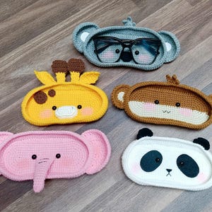 May include: Five crocheted animal-shaped trays. The trays are in the shapes of a koala, giraffe, monkey, elephant, and panda. The trays are all different colors and have cute faces.