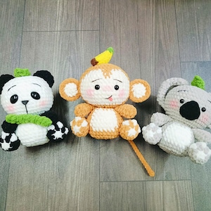 May include: Three crocheted stuffed toys: a panda with black and white fur, a monkey with brown fur and a banana on its head, and a koala with grey fur. The toys are all smiling and have big eyes.