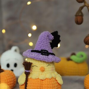 May include: A handmade crochet Halloween chick wearing a purple witch hat with a black buckle and bat accent. The chick is yellow and orange, holding a small broom. Other Halloween-themed crochet items are in the background.