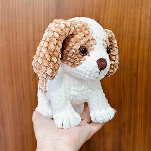 Puede incluir: Un perro de peluche tejido a crochet con pelo marrón y blanco. El perro tiene grandes ojos marrones y una pequeña nariz marrón.