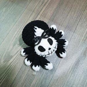 May include: A black and white crocheted spider with a skull face and a black ribbon tied around its head.