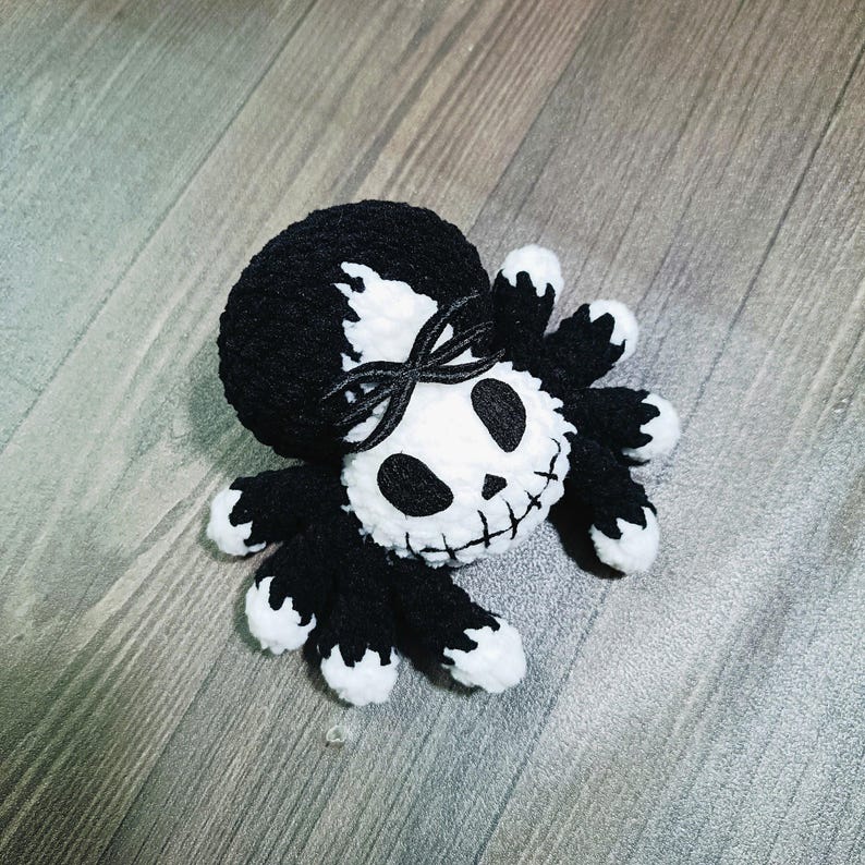May include: A black and white crocheted spider with a skull face and a black ribbon tied around its head.