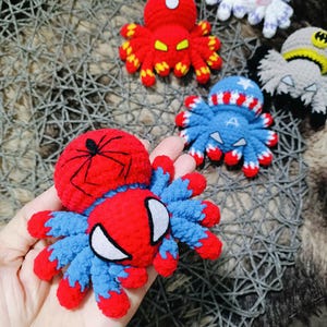 May include: Five crocheted spider toys, each with a different superhero theme. The spiders are red, blue, white, gray, and purple. The red spider has a black spider web on its back and a white spider face. The blue spider has a red, white, and blue pattern on its back and a white star on its head. The white spider has a purple pattern on its back. The gray spider has a black mask on its head. The purple spider has a white pattern on its back.