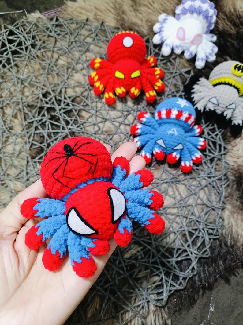 May include: Five crocheted spider toys, each with a different superhero theme. The spiders are red, blue, white, gray, and purple. The red spider has a black spider web on its back and a white spider face. The blue spider has a red, white, and blue pattern on its back and a white star on its head. The white spider has a purple pattern on its back. The gray spider has a black mask on its head. The purple spider has a white pattern on its back.