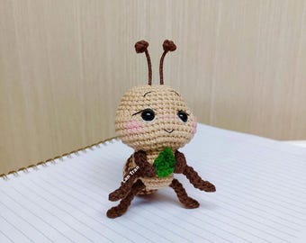 Può includere: Una bambola di formica all'uncinetto con testa e corpo beige, zampe e antenne marroni e una foglia verde. La bambola ha occhi neri e guance rosa. La bambola è seduta su un quaderno bianco.
