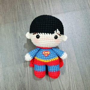 May include: A crocheted toy of a superhero wearing a red, blue, and yellow costume with a yellow 'S' on the chest. The toy has black hair and a white face with black eyes.