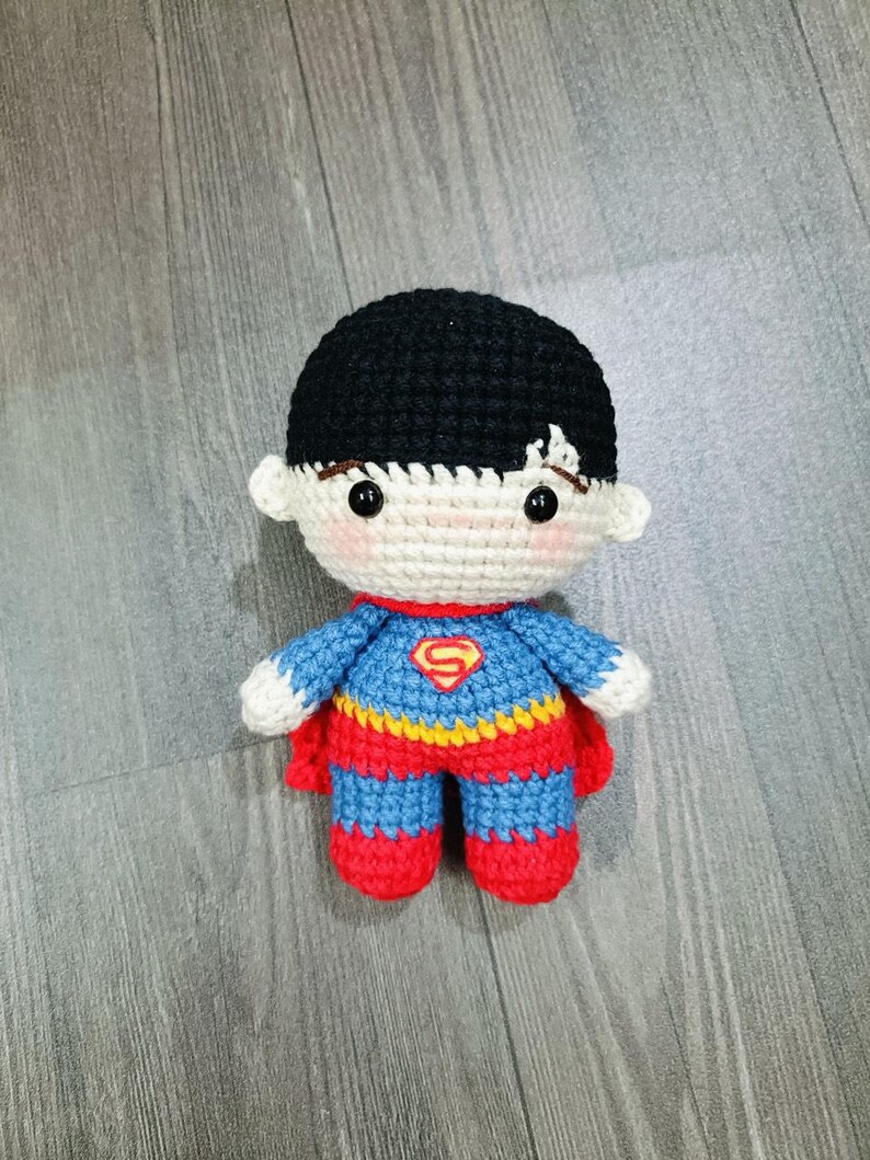 May include: A crocheted toy of a superhero wearing a red, blue, and yellow costume with a yellow 'S' on the chest. The toy has black hair and a white face with black eyes.
