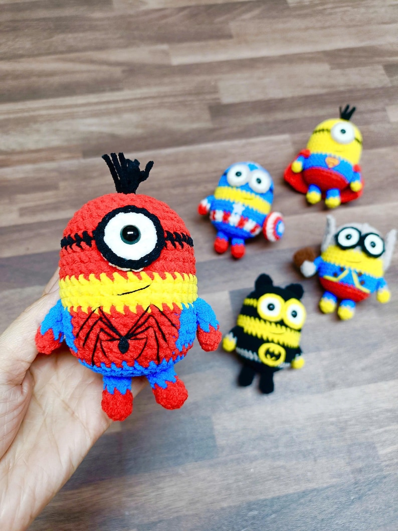 May include: A collection of handmade crochet Minion figures, each designed as a different superhero. The figures are primarily yellow, blue, red, and black, with details like a spider web and a Captain America shield.