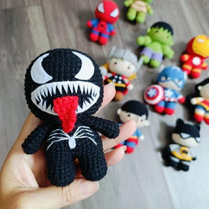 May include: A black crocheted Venom character with a red tongue and white eyes. The character is standing on a hand.