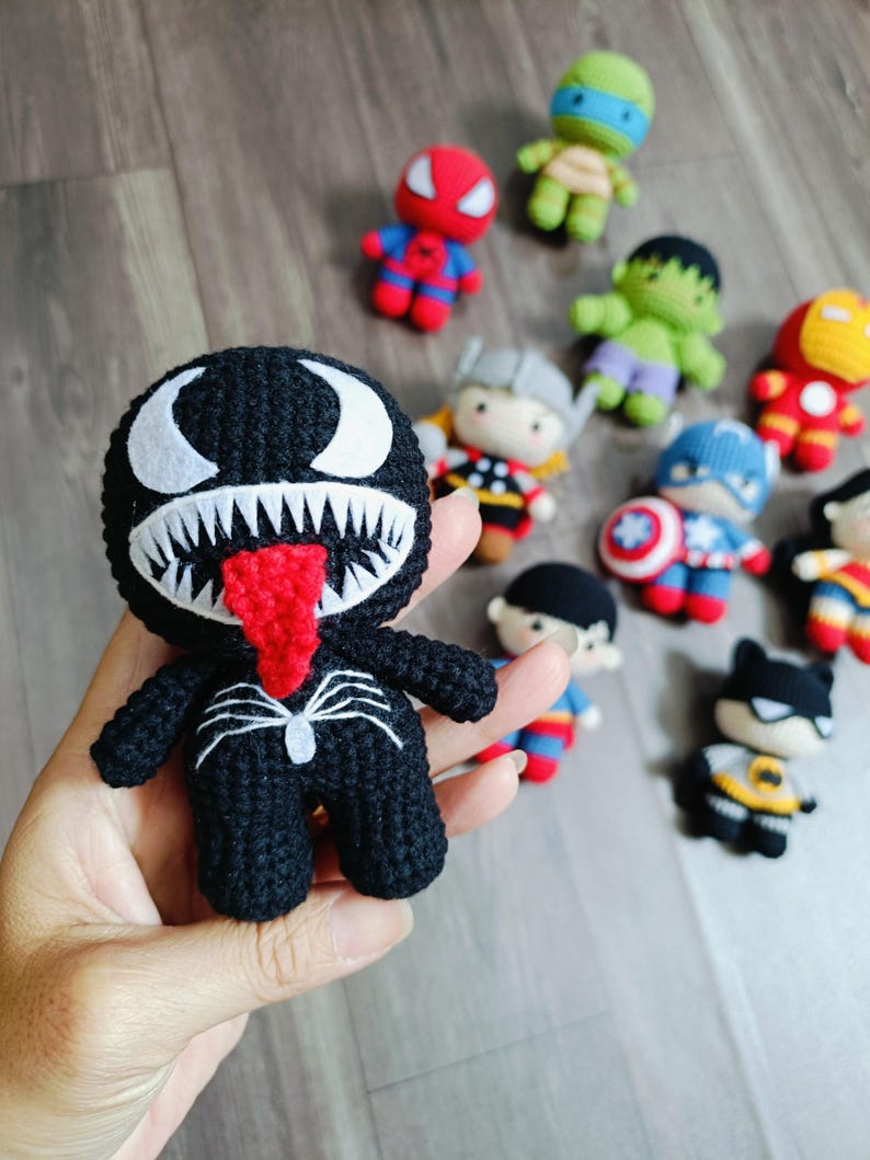 May include: A black crocheted Venom character with a red tongue and white eyes. The character is standing on a hand.