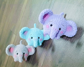 May include: Three crocheted elephant plushies in different colors: purple, blue, and gray. Each plush has pink ears and a small, embroidered face.