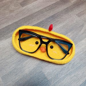 May include: A yellow crocheted glasses case shaped like a chick with black glasses inside. The chick has a red beak and black eyes.
