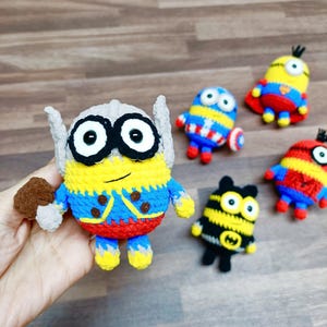 May include: A collection of handmade crochet Minion figures, each dressed as a superhero. The main figure is Thor, with a hammer. Other figures include Captain America, Superman, and Batman. The figures are yellow, blue, red, and black.