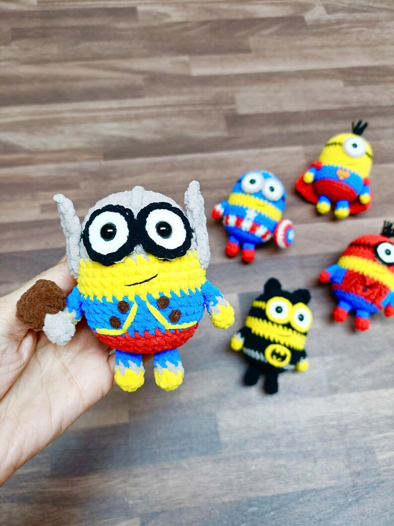 May include: A collection of handmade crochet Minion figures, each dressed as a superhero. The main figure is Thor, with a hammer. Other figures include Captain America, Superman, and Batman. The figures are yellow, blue, red, and black.