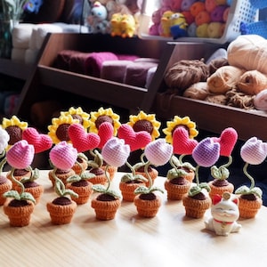 Może przedstawiać: Kolekcja szydełkowych aranżacji kwiatowych w doniczkach. Kwiaty są wykonane z różowego, fioletowego i białego włóczki. Doniczki są brązowe i mają zielone liście z włóczki. Jest mała szydełkowa słonecznik z czerwonym sercem w środku. Mała szydełkowa figurka kota znajduje się na pierwszym planie.