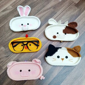 May include: Five crocheted animal-shaped eyeglass holders. The holders are in the shapes of a rabbit, cow, chicken, cat, and pig. The holders are all white with different colored accents.