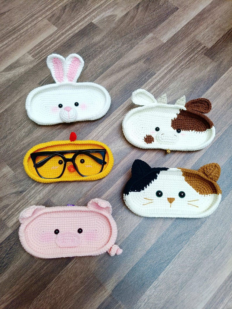 May include: Five crocheted animal-shaped eyeglass holders. The holders are in the shapes of a rabbit, cow, chicken, cat, and pig. The holders are all white with different colored accents.