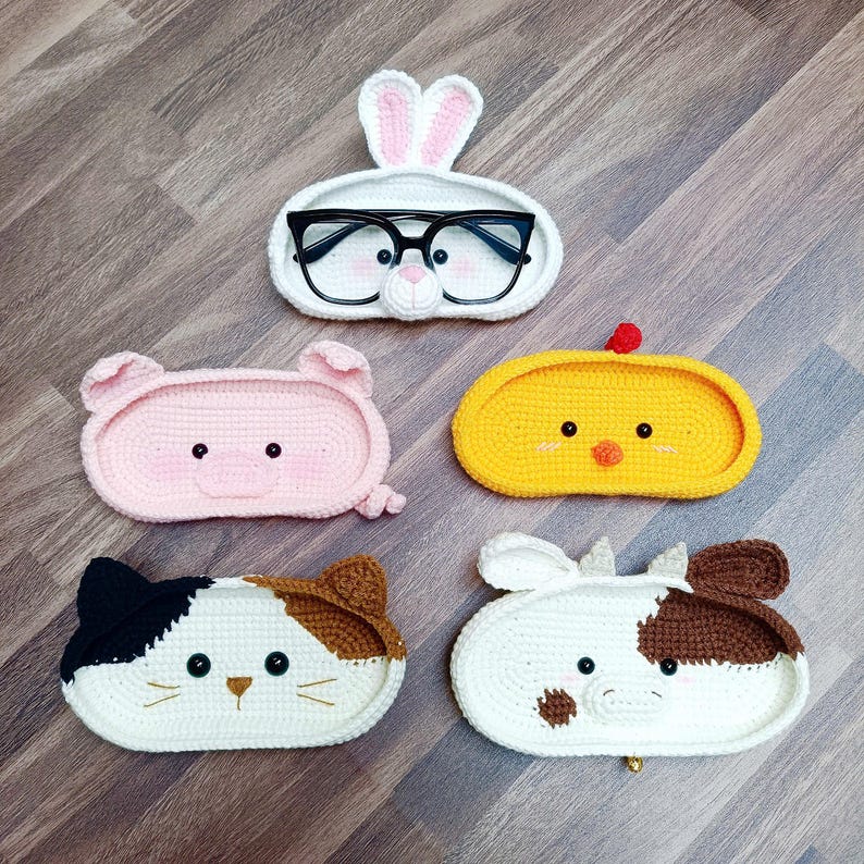 May include: Five crocheted animal-shaped eyeglass holders. The holders are shaped like a bunny, pig, chick, cat, and cow. Each holder has a cute face with embroidered eyes and a nose.