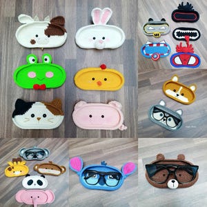 May include: A collection of crocheted eyeglass holders featuring various animal and cartoon characters. The holders are made in a variety of colors, including blue, green, pink, yellow, and brown. Each holder has a unique design, such as a frog, a pig, a bunny, a cow, a chicken, a fox, a cat, a panda, a Stitch character, and a bear.