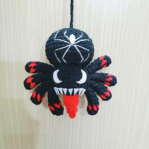 May include: A black and red crocheted spider with a white spider web design on its head. The spider has a red tongue sticking out.
