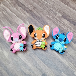 May include: Three handmade crochet plush toys in pink, orange, and blue. Each toy has large ears, black eyes, and a smaller matching toy. The toys are arranged on a wooden surface.