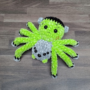 May include: A handmade Halloween-themed plush spider. The spider has a green body and legs, with a Frankenstein-inspired head featuring black hair, stitches, and a scar. The spider's abdomen is gray with black button eyes. The legs have black and gray accents.