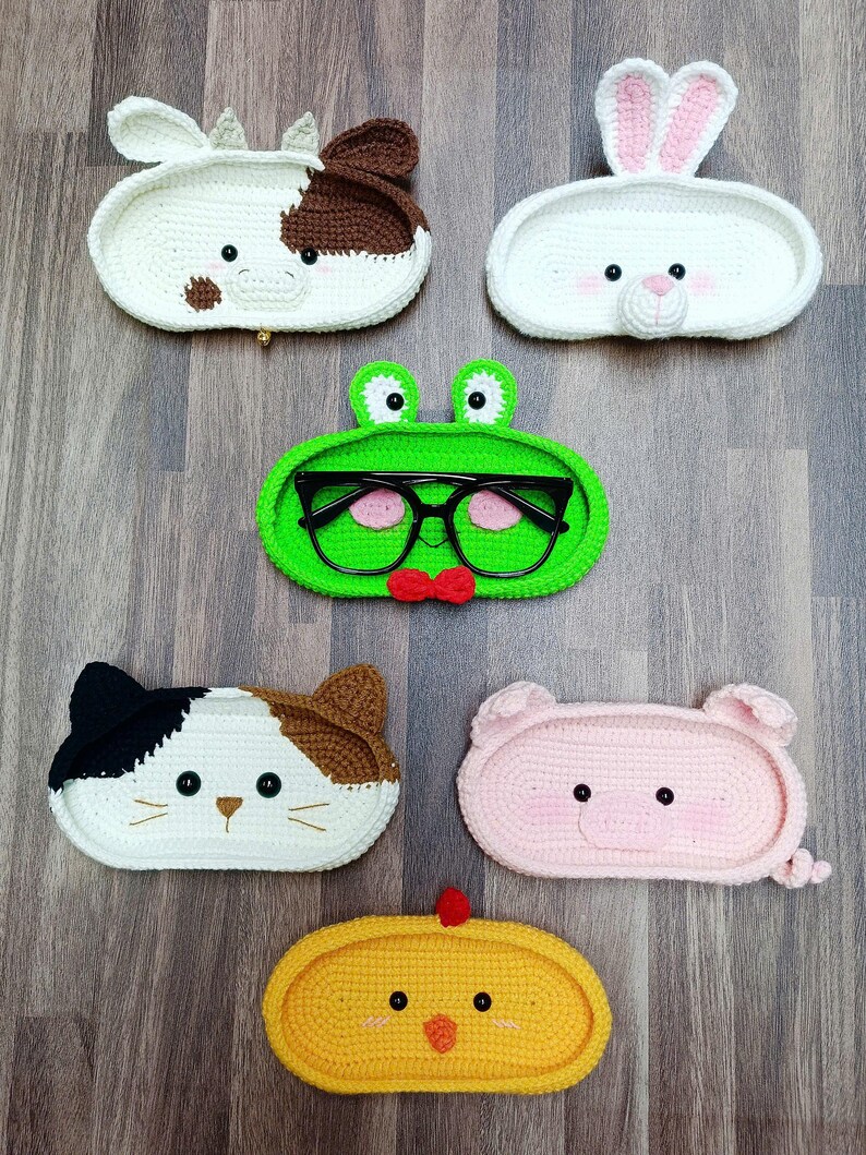 May include: Six crocheted animal-shaped trinket dishes. The dishes are shaped like a cow, rabbit, frog, cat, pig, and chick. Each dish has a cute face with embroidered details.